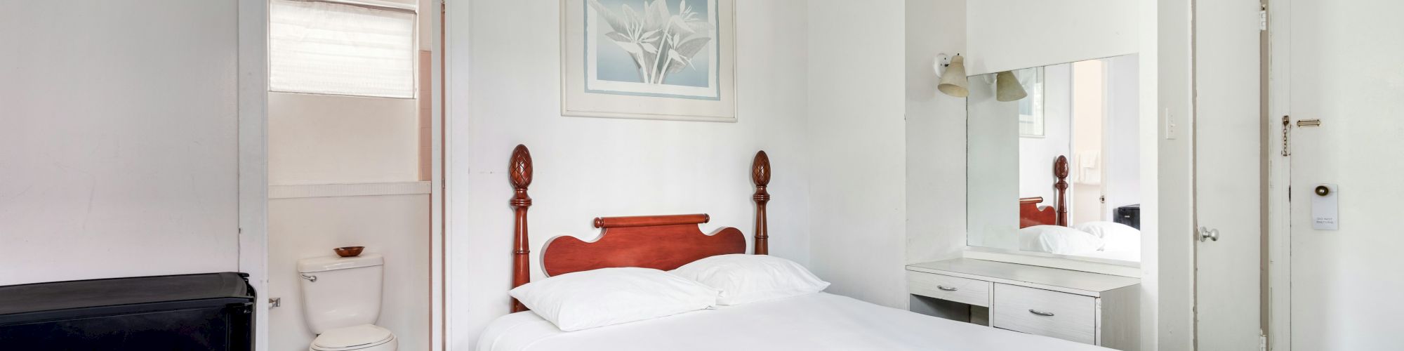 A bright bedroom with a double bed, mirror dresser, and an open doorway to a small bathroom with toilet and sink.
