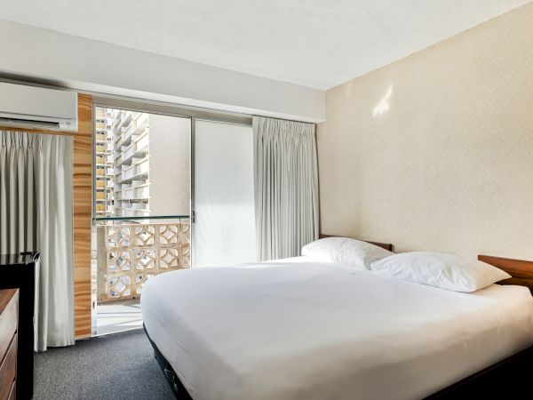 A modern hotel bedroom with a double bed, wall AC, curtains, and an open balcony door.
