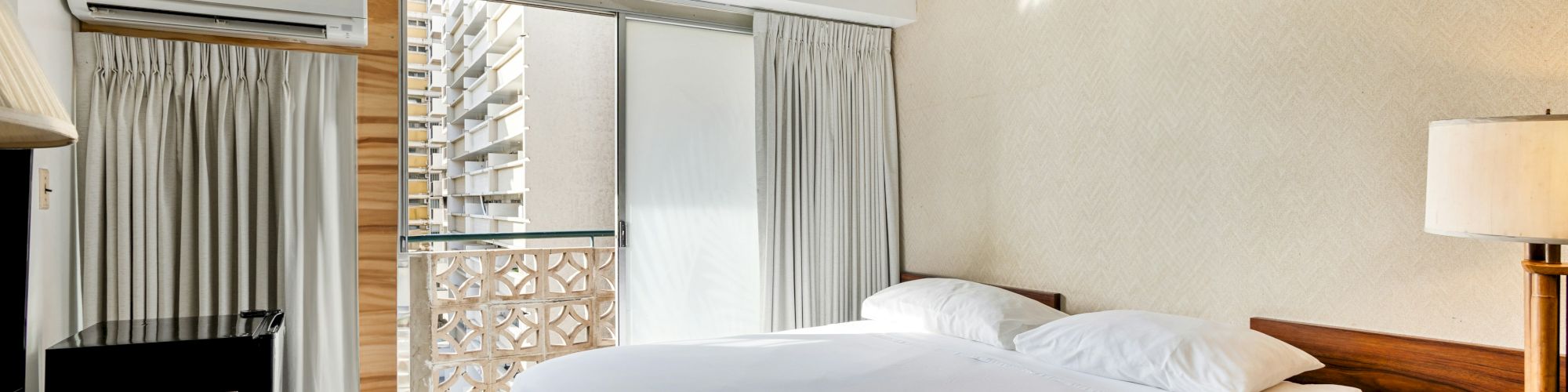 A modern hotel bedroom with a large bed, curtains, air conditioner, and an open balcony showing apartment buildings.