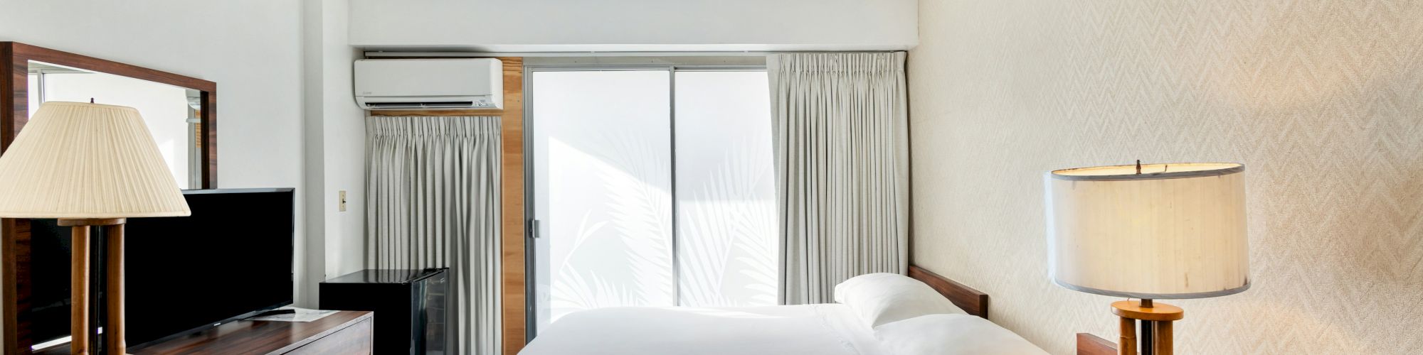 A tidy hotel-style bedroom with a large bed, bedside lamps, a TV, an air conditioner, and curtains over a sliding door.