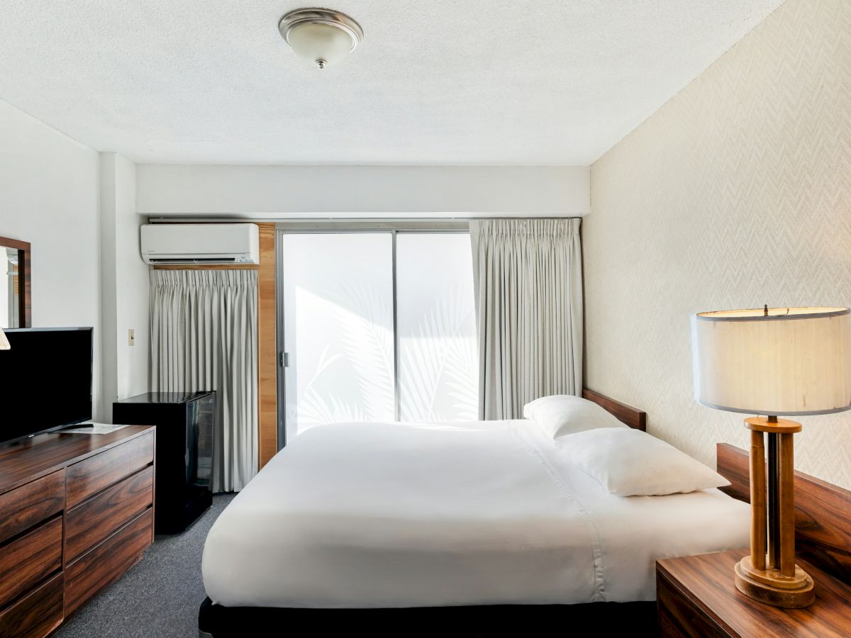 A tidy hotel bedroom with a queen bed, bedside lamps, TV on a dresser, and curtains by a balcony door with AC.
