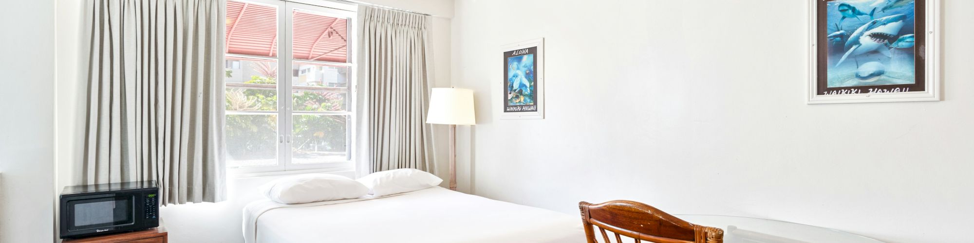 A bright small bedroom with a single bed, curtains and window, a lamp, framed ocean art, a microwave, and a round table with chairs.