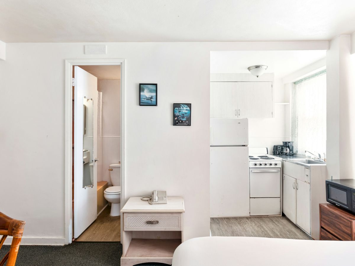 Bright apartment kitchenette with stove, sink, cabinets, microwave, and a doorway to a small bathroom.