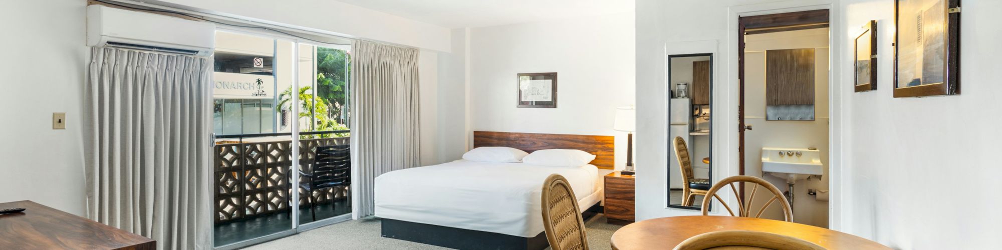 A bright hotel room with a bed, wooden dresser, rattan chairs at a table, and sliding glass doors with curtains.