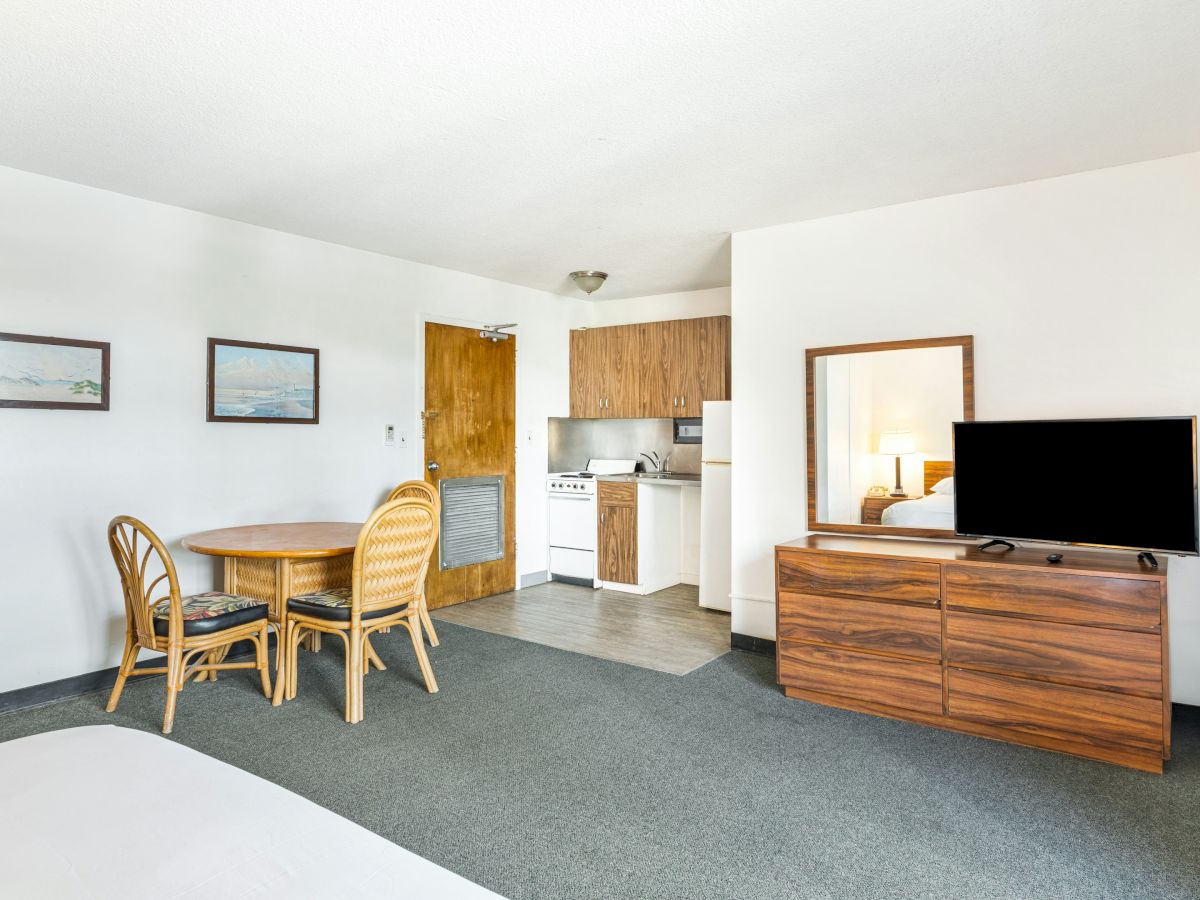 A hotel living room with a small kitchenette, dining table and chairs, a large TV on a wooden dresser, and framed wall art.