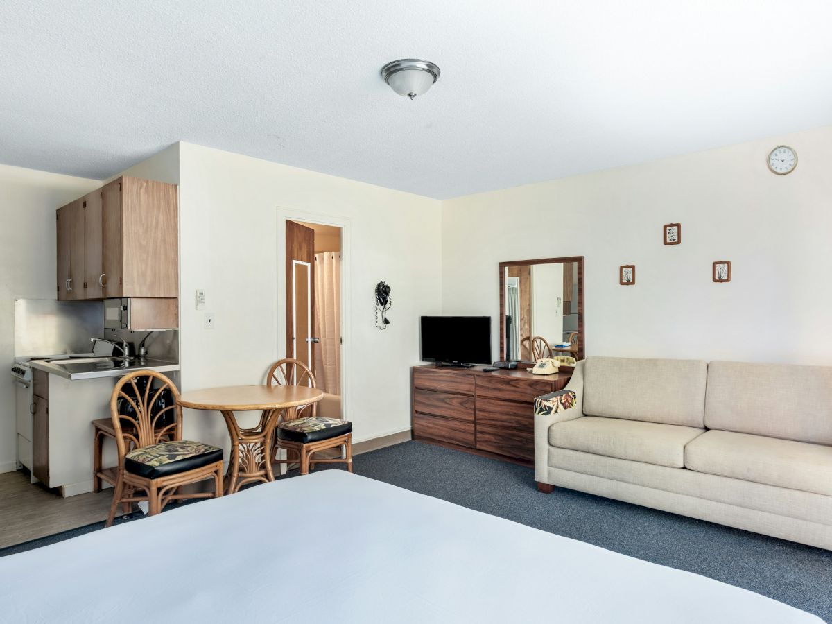 A bright living room with sofa, TV stand, dining table, kitchenette, chairs, and a ceiling clock.