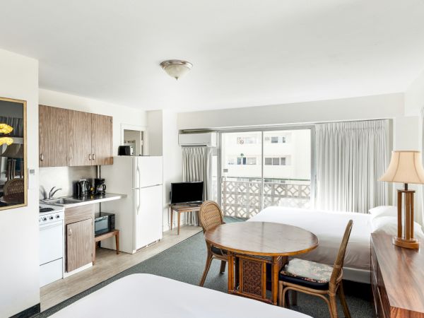 Bright hotel studio with kitchenette, round table, chairs, floor lamp, and large window with curtains.