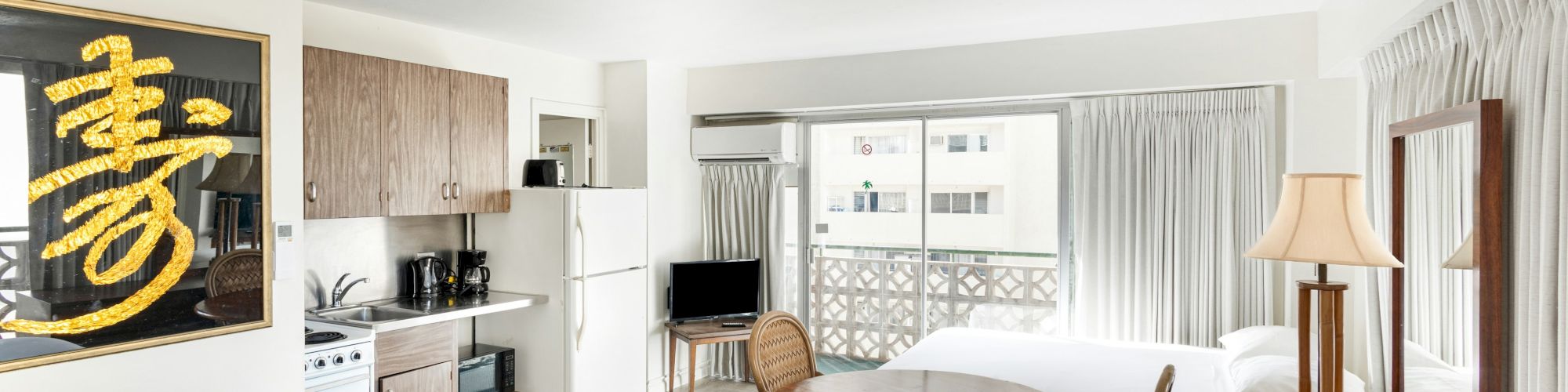 A bright studio apartment interior with a small kitchen, dining table, curtains, and lamps, plus a decorative gold wall sign.