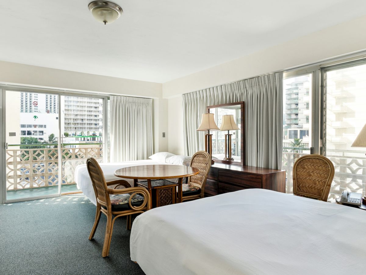 A bright hotel room with two beds, a wooden table and chairs, and large windows opening to a balcony with city views.