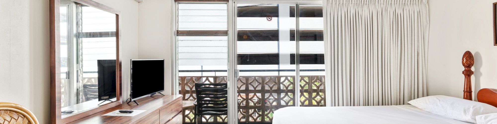 Hotel room with a bed, dresser and TV, sliding glass doors to a balcony, and curtains with an air conditioner above.