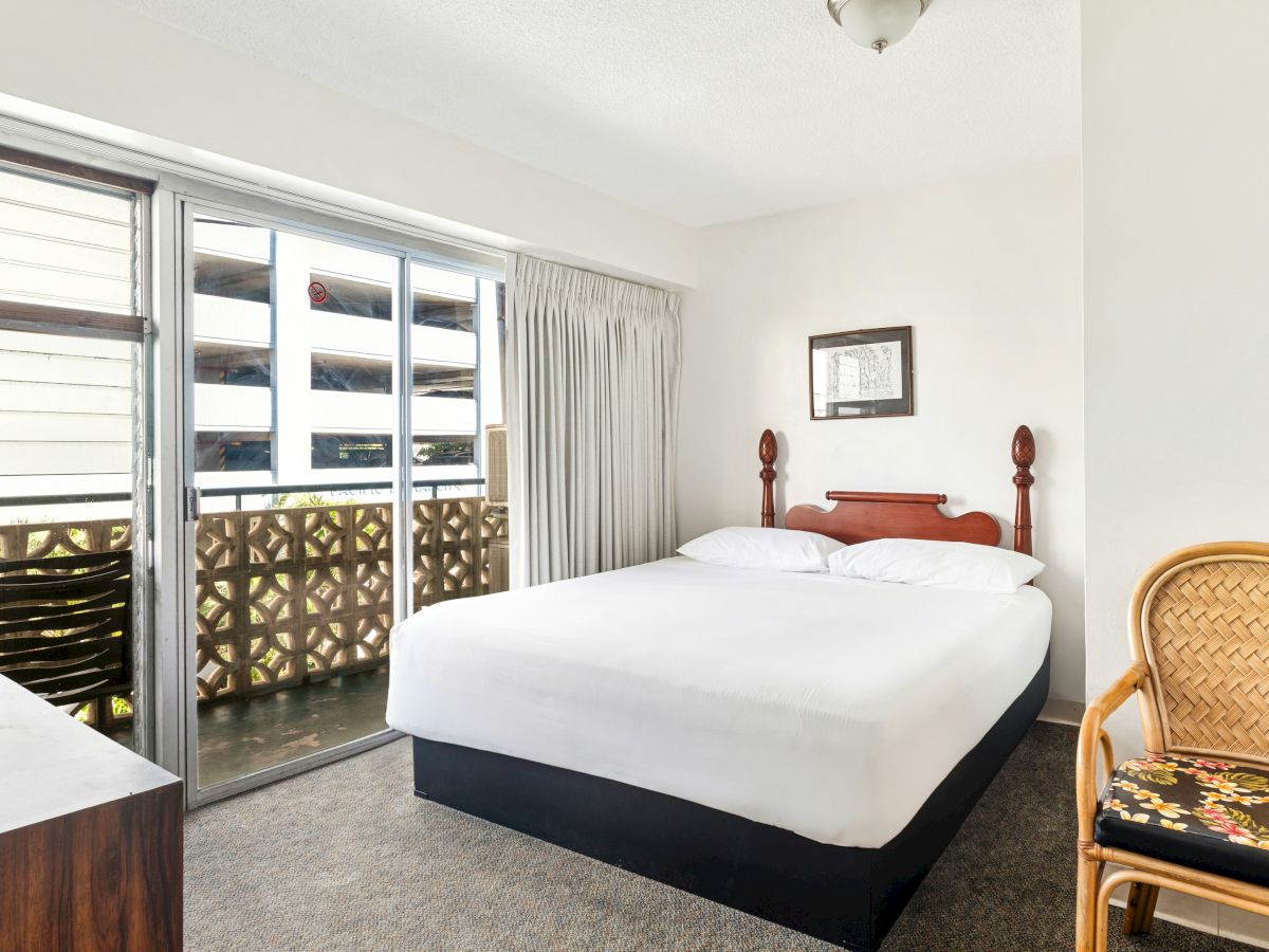 Bright bedroom with a wooden bed, white bedding, wicker chair, framed art, and large sliding doors opening to a balcony.