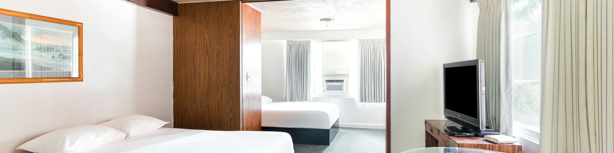 A bright hotel room with two beds, a TV on a stand, curtained windows, and a glass table.