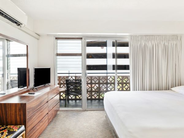 A bright hotel room with a bed, TV and wooden dresser, plus sliding balcony doors, curtains, and a wall air conditioner.