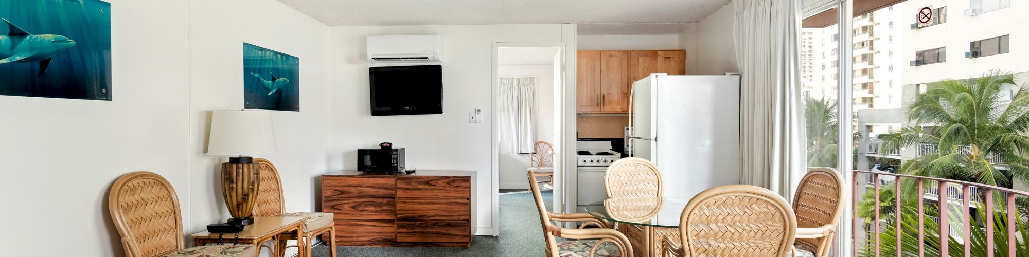 Bright studio room with wicker chairs, kitchenette, TV, and an open balcony view with palm plants outside.