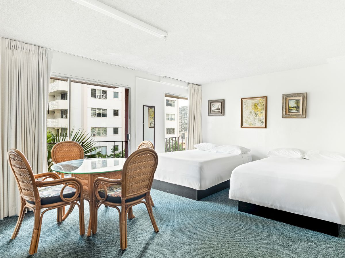 Bright hotel room with two white beds, rattan chairs and a glass table, plus framed art and a view through open balcony doors.