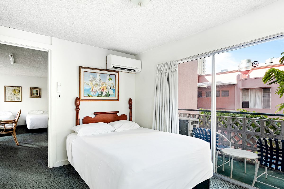A bright hotel bedroom with a large white bed, air conditioner, and a balcony view with plants and outdoor seating.