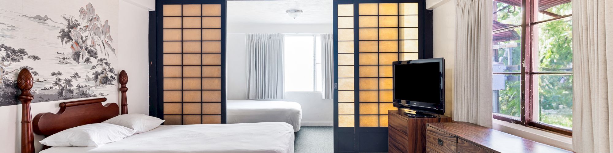 A modern hotel bedroom with a double bed, scenic mural, TV on a wooden dresser, and curtains by a window.