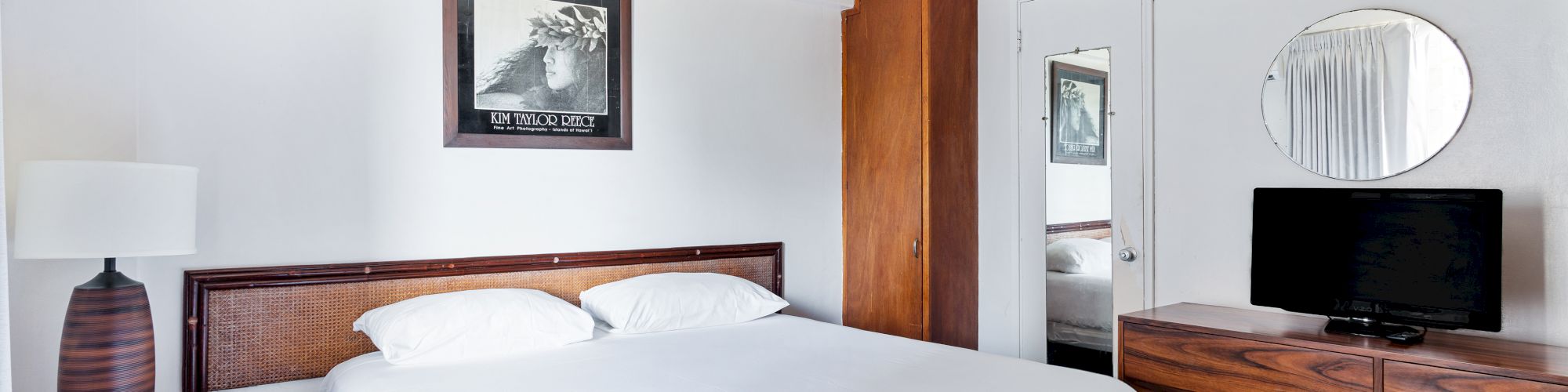 A neatly made bedroom with a double bed, wooden dresser, wall mirror, TV, and an open wardrobe with a second room visible.