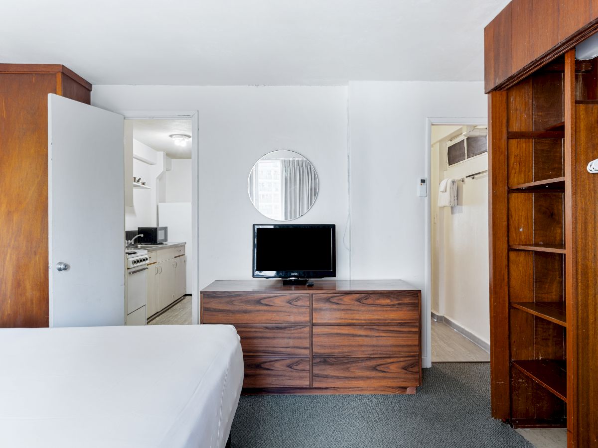 A tidy hotel room with a bed, TV on a wooden dresser, round mirror, kitchenette behind, and a closet with hangers.