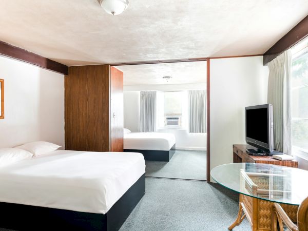 A bright hotel room with two beds, a TV on a stand, curtains, a window, and a small glass-topped table.