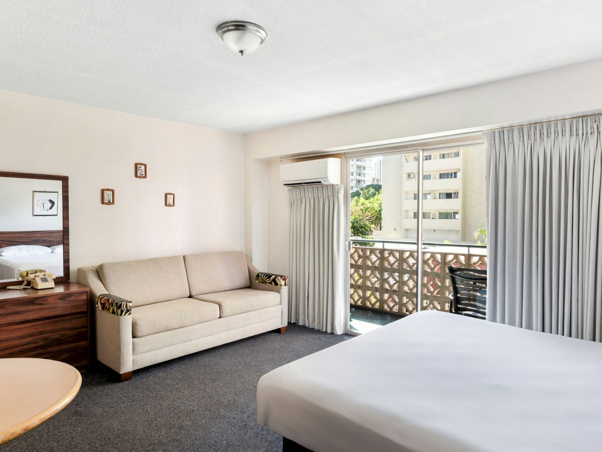 A bright hotel room with a bed, couch, wooden dresser, phone, window air conditioner, and gray curtains opening to a balcony.